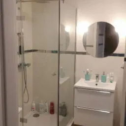 Nowoczesna łazienka z kabiną prysznicową typu walk-in, białą umywalką z szafką podwieszaną, okrągłym lustrem z podświetleniem i drewnianą podłogą.