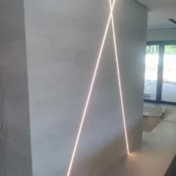 Ściana wyłożona jasnymi płytkami z podświetleniem LED tworzącym wzór krzyża, widoczne połączenie płytek na krawędziach, w tle przeszklone drzwi balkonowe.