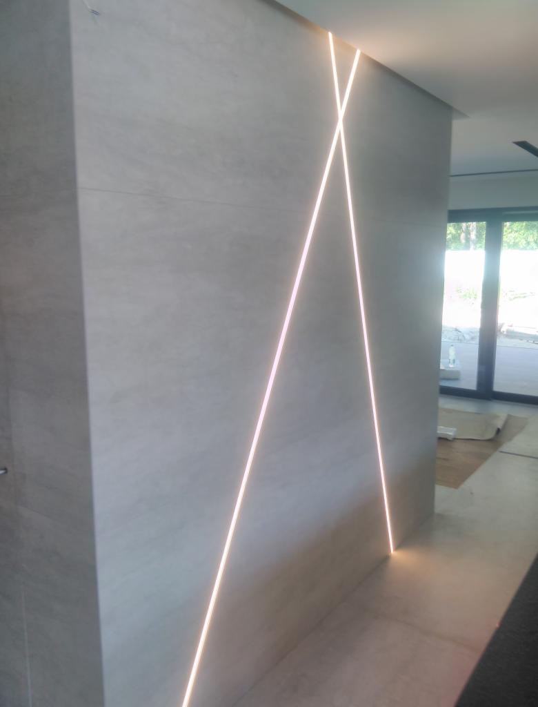 Ściana wyłożona jasnymi płytkami z podświetleniem LED tworzącym wzór krzyża, widoczne połączenie płytek na krawędziach, w tle przeszklone drzwi balkonowe.