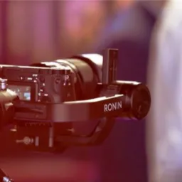 Profesjonalny aparat z gimbalem Ronin ustawiony w tle z rozmytą postacią w białej koszuli.