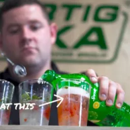 Barman w czarnej koszuli przygotowuje trzy drinki z owocami i miętą, nalewając napój z zielonej butelki Sprite do jednego z nich, używając długiej łyżki barmańskiej.