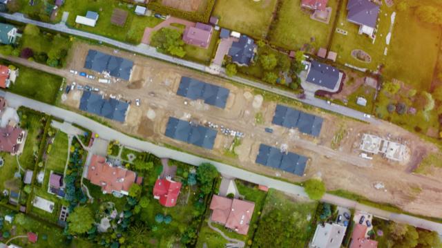 Widok z drona na plac budowy z kilkoma domami w stanie surowym zamkniętym i jednym domem w trakcie wznoszenia ścian, otoczony zielenią i istniejącą zabudową.