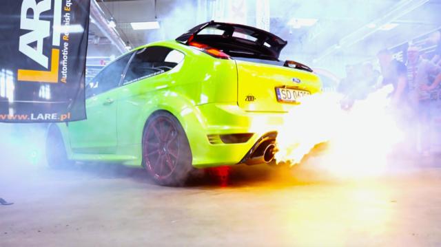 Tuningowany, jasnozielony Ford Focus RS z otwartą klapą bagażnika, z wydechu wydobywają się płomienie, czerwone felgi, widoczny baner reklamowy Lare
