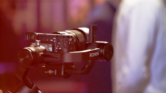 Profesjonalny aparat z gimbalem Ronin ustawiony w tle z rozmytą postacią w białej koszuli.