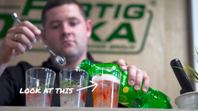 Barman w czarnej koszuli przygotowuje trzy drinki z owocami i miętą, nalewając napój z zielonej butelki Sprite do jednego z nich, używając długiej łyżki barmańskiej.