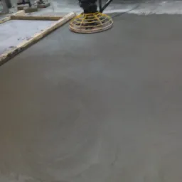 Zacieranie świeżej wylewki betonowej za pomocą zacieraczki mechanicznej z żółtą osłoną, widoczne drewniane szalunki i wózek widłowy w tle.