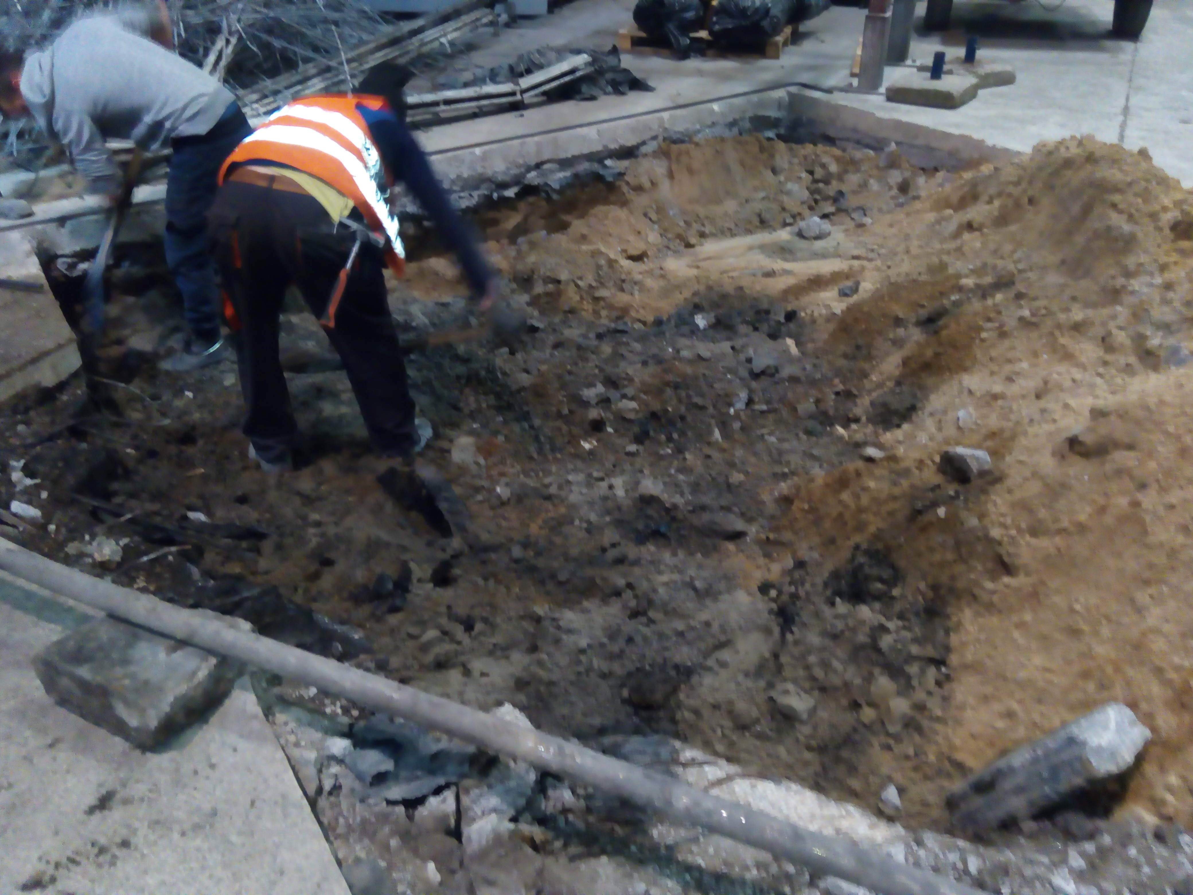 Dwóch pracowników w kamizelkach odblaskowych kopie dół w ziemi, widoczne narzędzia i wykopana ziemia. Prace ziemne wewnątrz budynku.