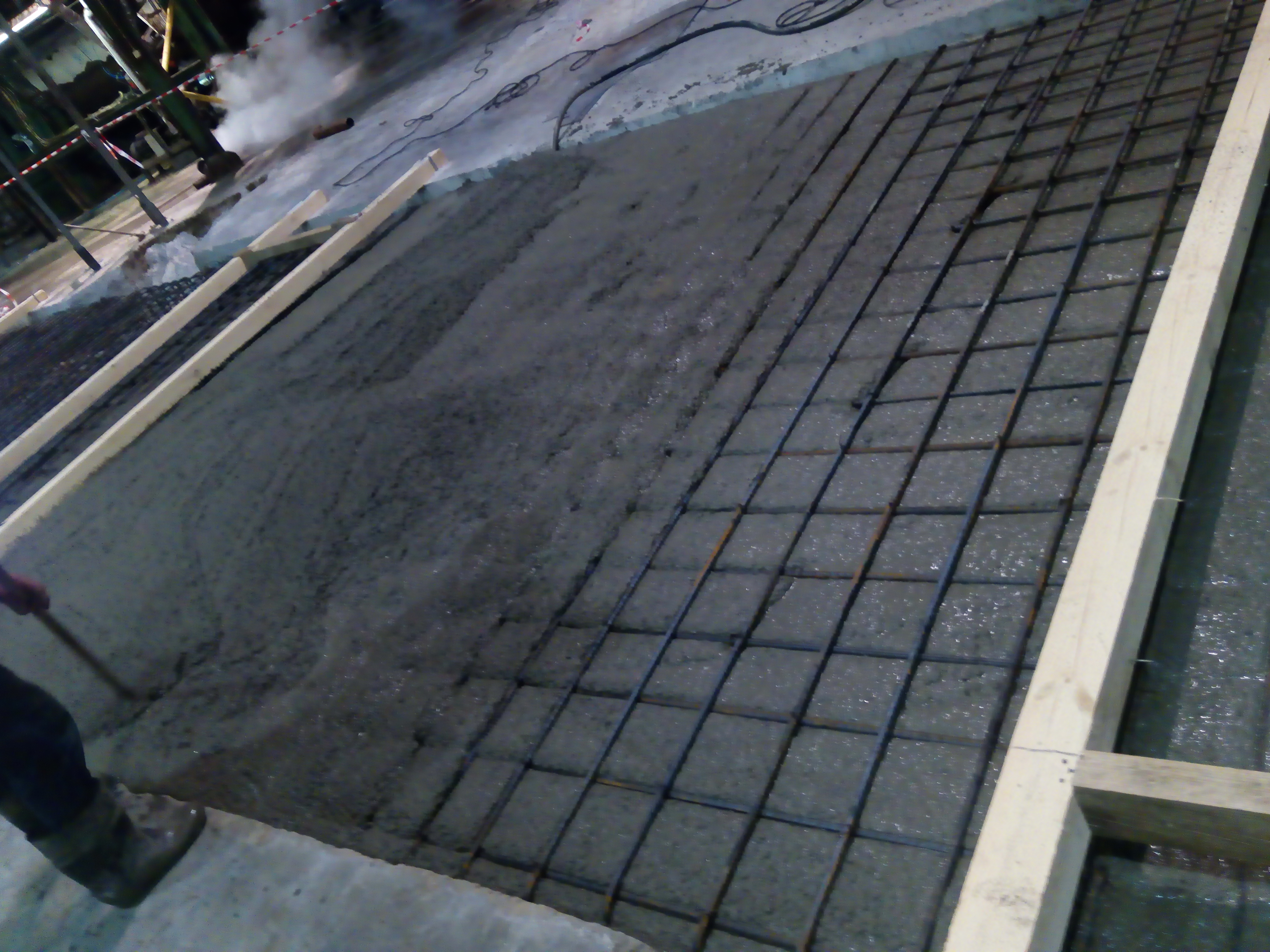 Świeżo wylana, mokra wylewka z siatką zbrojeniową, drewniane szalunki, widoczny pracownik w roboczych butach, w tle instalacje przemysłowe i para.