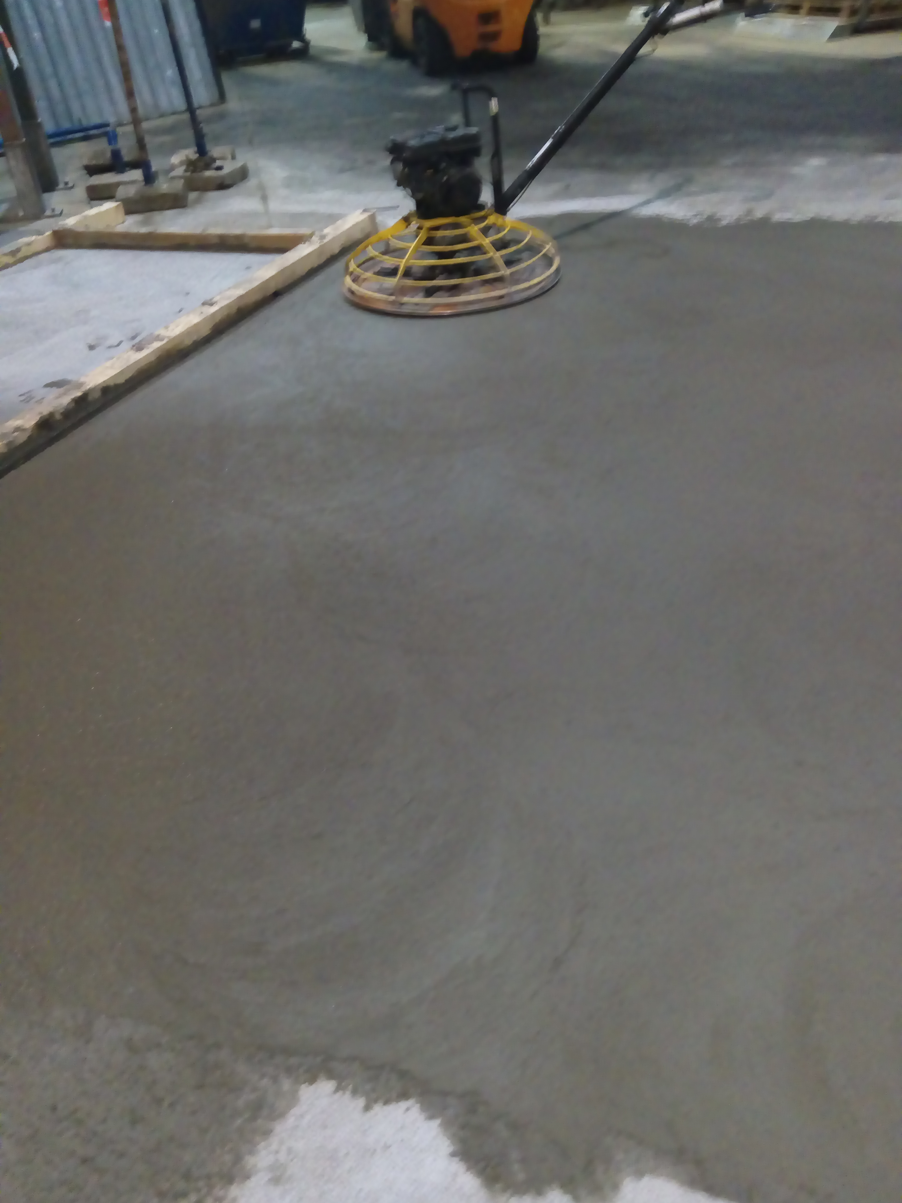 Zacieranie świeżej wylewki betonowej za pomocą zacieraczki mechanicznej z żółtą osłoną, widoczne drewniane szalunki i wózek widłowy w tle.