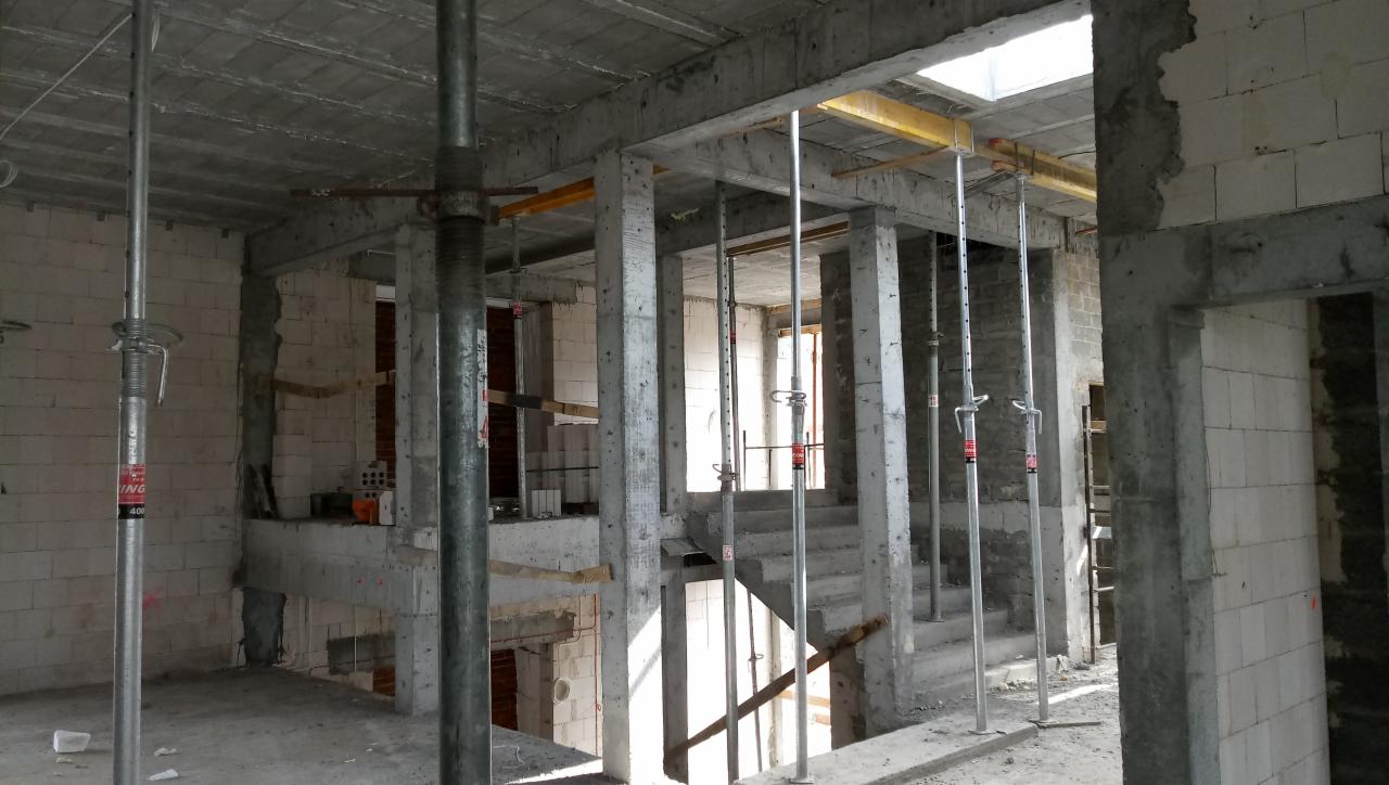 Wnętrze budynku w trakcie budowy, widoczne betonowe filary podtrzymujące strop, metalowe stemple budowlane, schody w surowym stanie i ściany z białych bloczków.