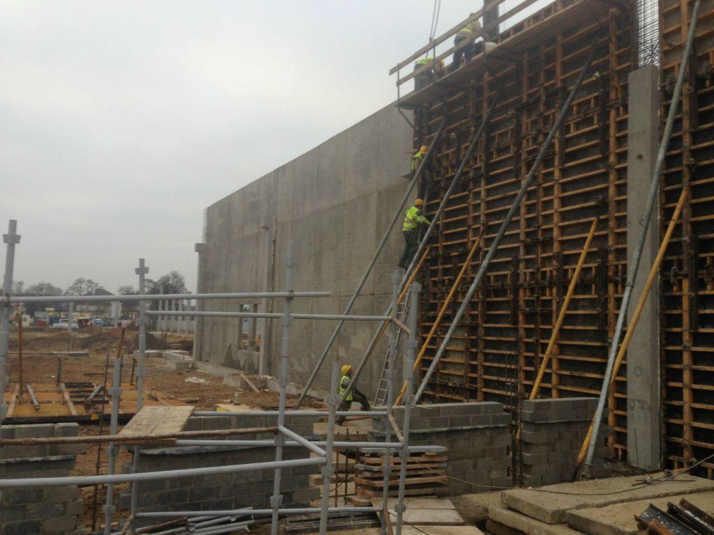 Wznoszenie betonowej ściany szalunkowej z widocznymi podporami i pracownikami w żółtych kamizelkach, na tle pochmurnego nieba i placu budowy z dźwigami.