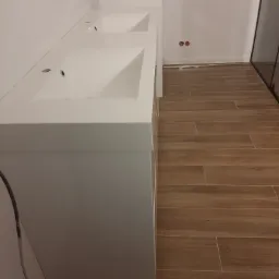 Nowoczesna, biała umywalka podwójna z szafką, zamontowana na tle drewnopodobnych płytek podłogowych w łazience.