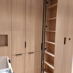 Zbigniew Czerech - Jasne, drewniane meble kuchenne z czarnymi uchwytami; otwarta szafka z półkami i czarną ramą drzwi. Widoczny fragment szuflady z mechanizmem.