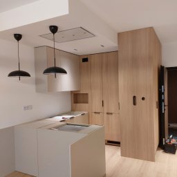 Zbigniew Czerech - Montaż jasnej kuchni z wyspą i drewnianymi szafami. Widoczne czarne lampy sufitowe i narzędzia w tle. Etap prac wykończeniowych.