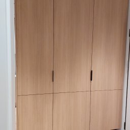 Zbigniew Czerech - Jasna, drewniana szafa wnękowa z minimalistycznymi, czarnymi uchwytami, stojąca na niskich nóżkach. Widok z perspektywy lekko z dołu, ukazujący fakturę drewna.