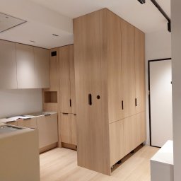 Zbigniew Czerech - Jasna zabudowa kuchenna z drewnianymi frontami i szafą wnękową, minimalistyczne uchwyty, oświetlenie punktowe na suficie, drewniana podłoga.