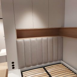 Zbigniew Czerech - Nowoczesna sypialnia z beżowym tapicerowanym zagłówkiem, drewnianą półką i szafkami w zabudowie. Widoczny stelaż łóżka i oświetlenie.