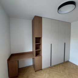 Zbigniew Czerech - Jasna szafa wnękowa z długimi, czarnymi uchwytami i biurko z drewnianym blatem, połączone w jedną całość. Podłoga w jodełkę, nowoczesna lampa sufitowa.