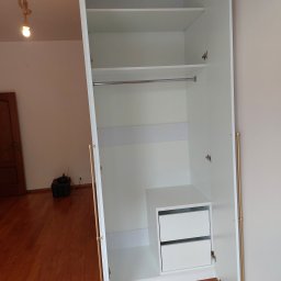 Zbigniew Czerech - Wnętrze białej szafy wnękowej z drążkiem na ubrania i szufladami. Widoczne okucia w kolorze złotym. Na podłodze drewniany parkiet i sprzęt elektroniczny.