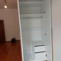 Wnętrze białej szafy wnękowej z drążkiem na ubrania i szufladami. Widoczne okucia w kolorze złotym. Na podłodze drewniany parkiet i sprzęt elektroniczny.