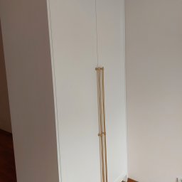 Zbigniew Czerech - Biała szafa dwudrzwiowa z długimi, złotymi uchwytami, umieszczona przy jasnej ścianie. Widoczny fragment podłogi. Minimalistyczny design.