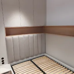 Kompletne łóżko z tapicerowanym wezgłowiem, szafką nocną i szafkami wiszącymi w jasnej kolorystyce. Widoczny stelaż łóżka i drewniana półka nad wezgłowiem.