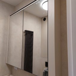 Zbigniew Czerech - Lustrzana szafka łazienkowa z czarnymi detalami, odbicie grzejnika drabinkowego. Minimalistyczny design, kremowe płytki na ścianie. Umywalka w trakcie montażu.