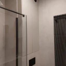 Nowoczesna łazienka z kabiną prysznicową typu walk-in, beżowymi płytkami i pionowym grzejnikiem w kolorze czarnym. Widoczna zabudowa meblowa w jasnym odcieniu.