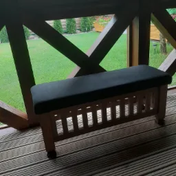 Drewniana ławka z ciemnoszarym siedziskiem na tarasie z drewnianą podłogą, widok na zielony trawnik i krzewy przez drewnianą konstrukcję zadaszenia.