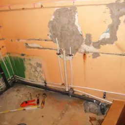 Wnętrze łazienki w trakcie remontu, widoczne nowe rury instalacji wodno-kanalizacyjnej mocowane do ściany, narzędzia na podłodze, ściany w stanie surowym z fragmentami starej farby i tynku.
