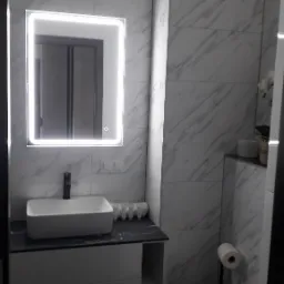 Nowoczesna łazienka z marmurowymi płytkami, podświetlanym lustrem i białą umywalką na szafce. Minimalistyczny wystrój z akcentami szarości i czerni.