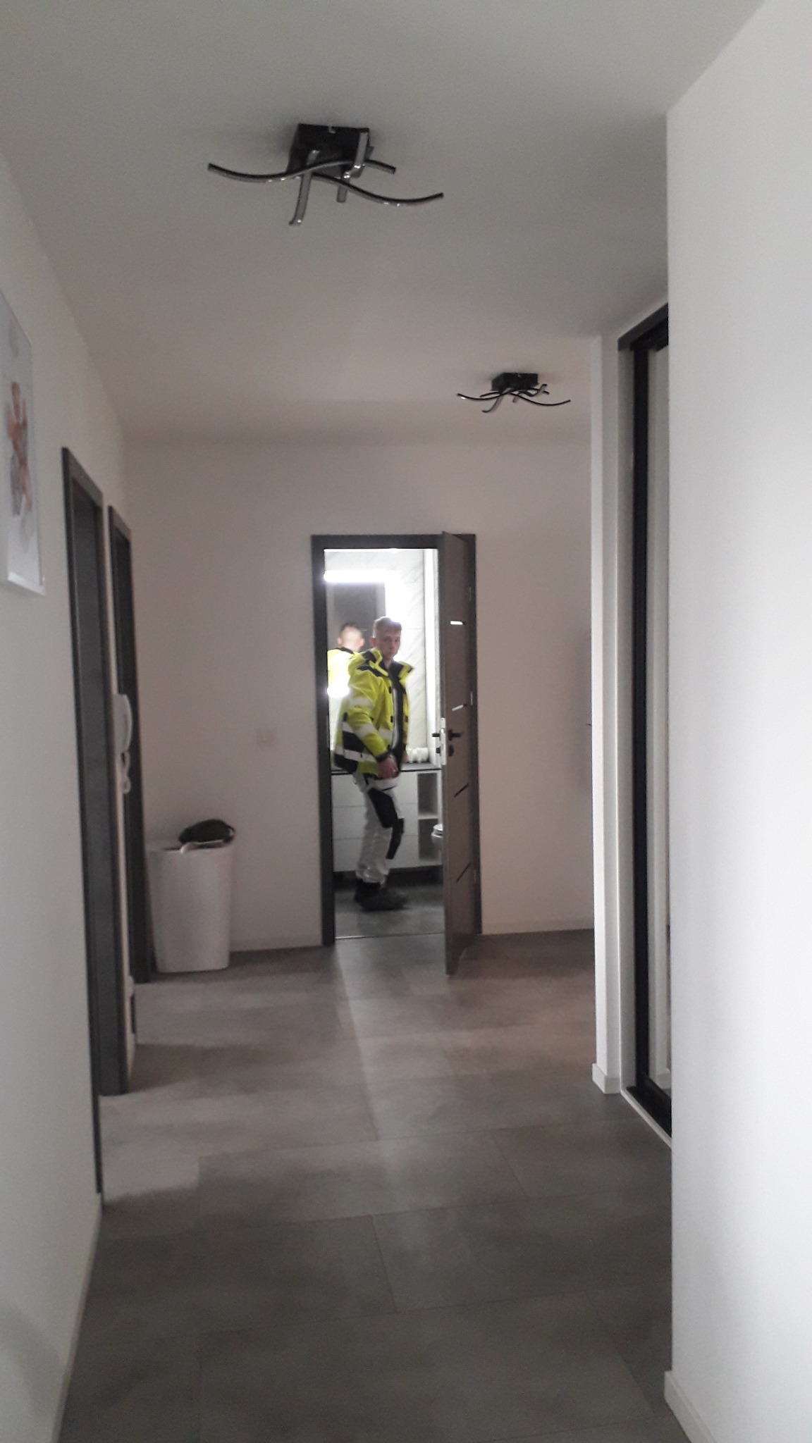 Korytarz z szarą podłogą i białymi ścianami. Widoczny mężczyzna w jaskrawej kurtce w otwartych drzwiach do łazienki. Na suficie dwie lampy.