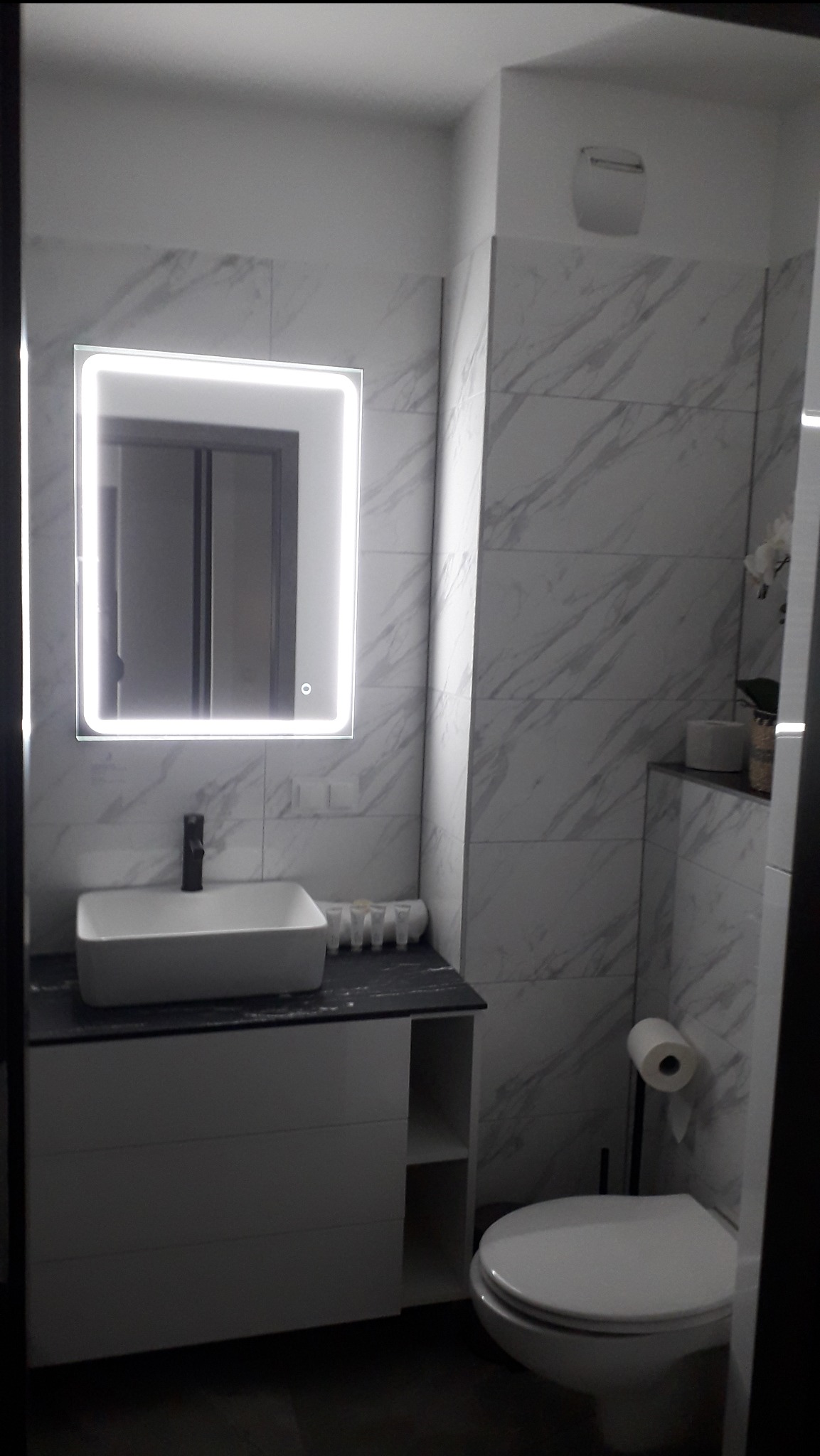 Nowoczesna łazienka z marmurowymi płytkami, podświetlanym lustrem i białą umywalką na szafce. Minimalistyczny wystrój z akcentami szarości i czerni.