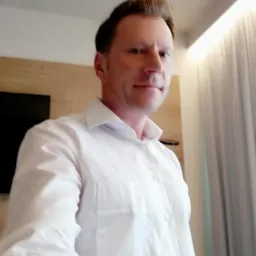 Portret mężczyzny w białej koszuli na tle pokoju hotelowego z drewnianą ścianą i zasłoną.