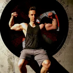 Młody mężczyzna w szarej koszulce na ramiączkach i krótkich spodenkach, siedzący w okrągłej wnęce z artystycznym wzorem, prezentuje napięte bicepsy, patrząc w obiektyw.