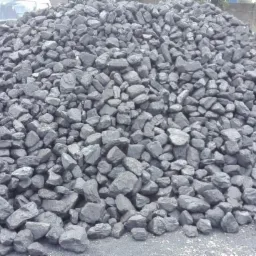 Góra czarnych bryłek węgla na placu składowym, z widocznym samochodem dostawczym w tle.