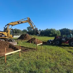 Żółta koparka CAT M312 i czerwony wózek teleskopowy Manitou pracują na zielonej łące przy wykopie pod fundamenty, widoczne drewniane znaczniki.