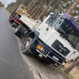 Ciężarówka MAN z naczepą przewożąca koparkę CAT na tle drzew, widok z przodu na poboczu drogi.