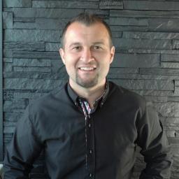 Moonfield Studio - Uśmiechnięty mężczyzna w czarnej koszuli na tle ściany z szarej, łupkowej okładziny. Widoczny fragment wzorzystej podszewki koszuli.