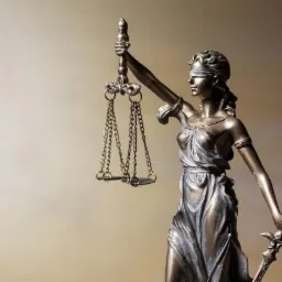 Brązowa statuetka Temidy z zasłoniętymi oczami trzymająca wagę i miecz na rozmytym beżowym tle.