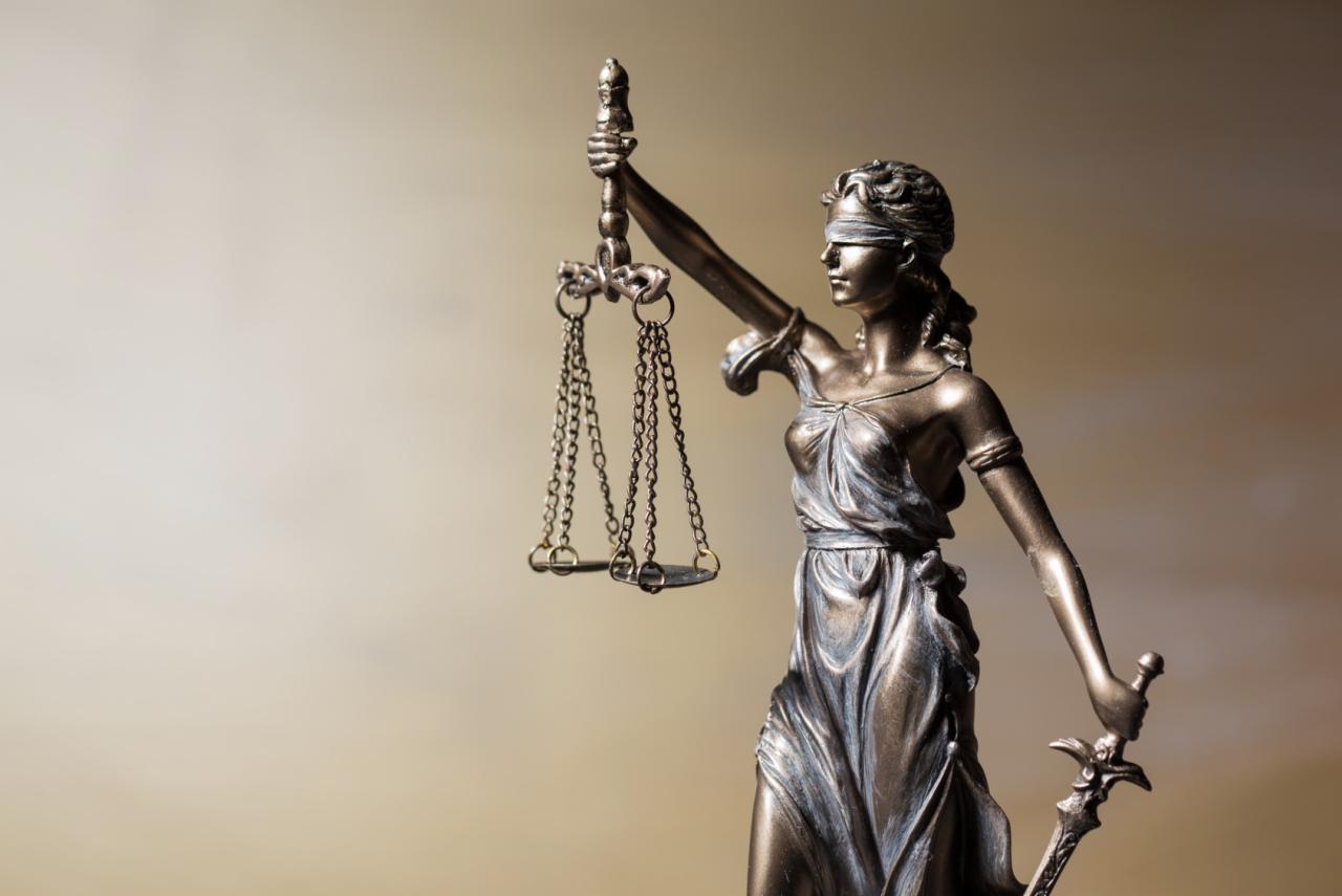 Brązowa statuetka Temidy z zasłoniętymi oczami trzymająca wagę i miecz na rozmytym beżowym tle.