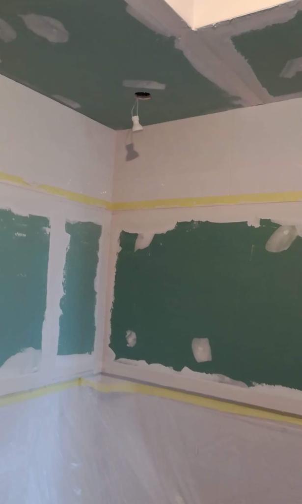 Pokój w trakcie szpachlowania: zielone ściany z widocznymi łatami szpachli, zabezpieczone taśmą malarską, podłoga przykryta folią ochronną, sufit z widocznymi łączeniami płyt karton-gips...