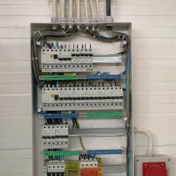 Otwarta skrzynka elektryczna z licznymi bezpiecznikami, przewodami różnej grubości i koloru, listwami zaciskowymi oraz dodatkowym gniazdem zewnętrznym z czerwonym i niebieskimi wtykami, zamontowana...