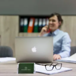 Biuro: Kobieta w błękitnej koszuli rozmawia przez telefon siedząc przy biurku z laptopem, wizytówką, okularami i stosem dokumentów; w tle regał z segregatorami.