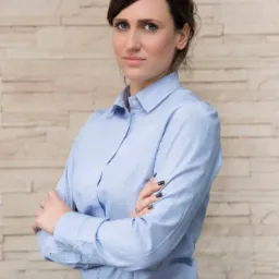 Portret kobiety w błękitnej koszuli z założonymi rękami na tle ściany z dekoracyjnego kamienia.