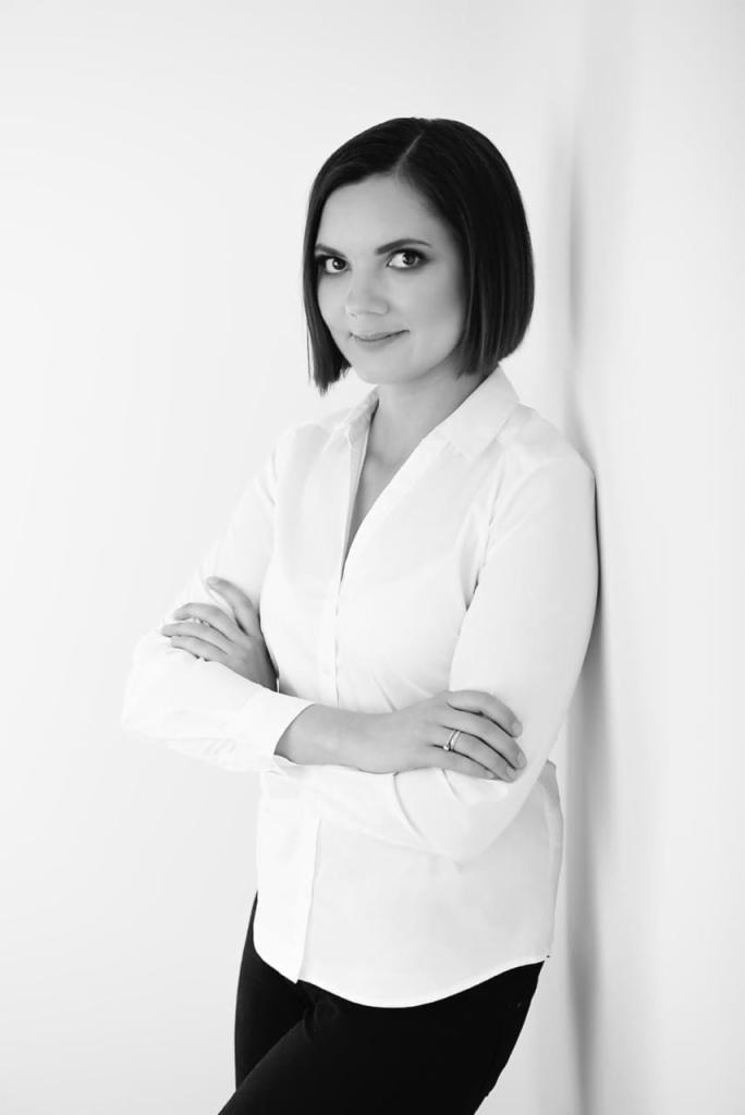 Portret kobiety w białej koszuli, opierającej się o białą ścianę, z założonymi rękami i subtelnym uśmiechem, w czarno-białej tonacji.