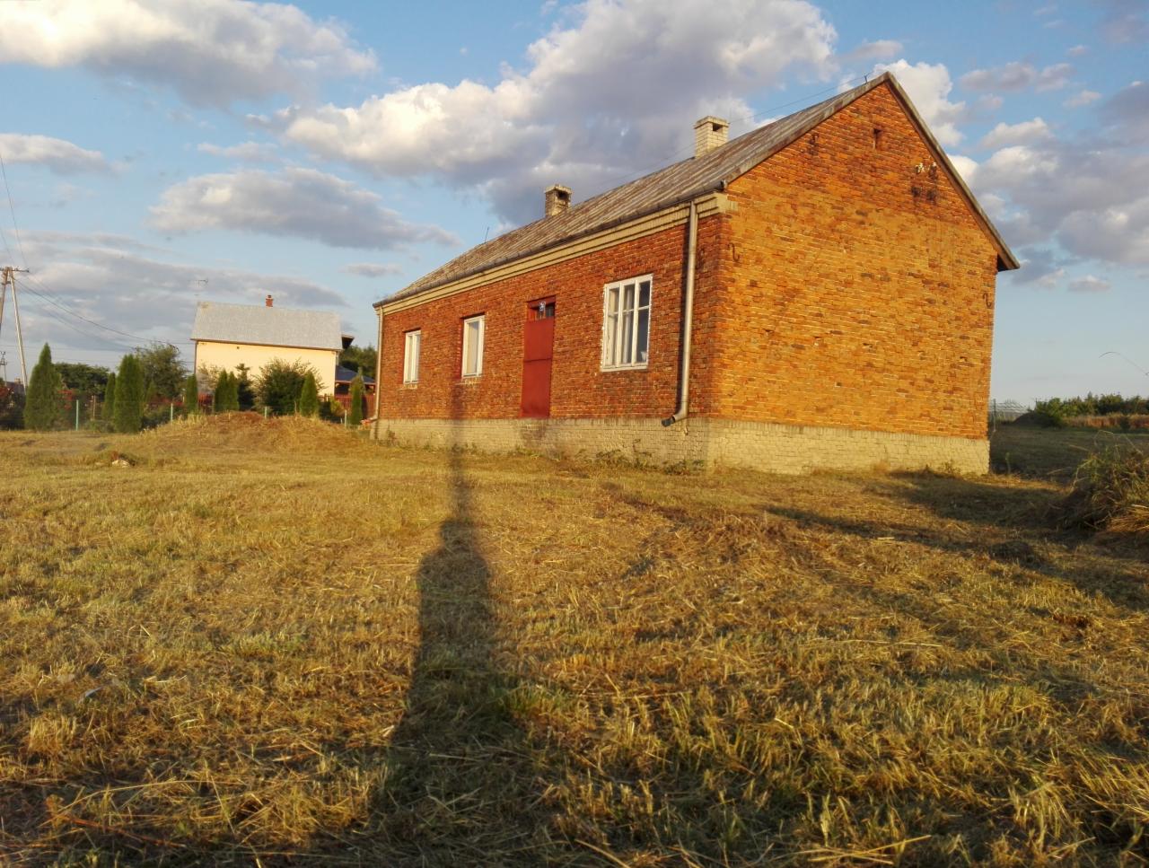 Czerwony ceglany dom z czerwoną stolarką okienną i drzwiami, otoczony suchą, żółtą trawą, z cieniem osoby rzucanym na pierwszy plan w słoneczny dzień z błękitnym niebem i białymi chmurami.