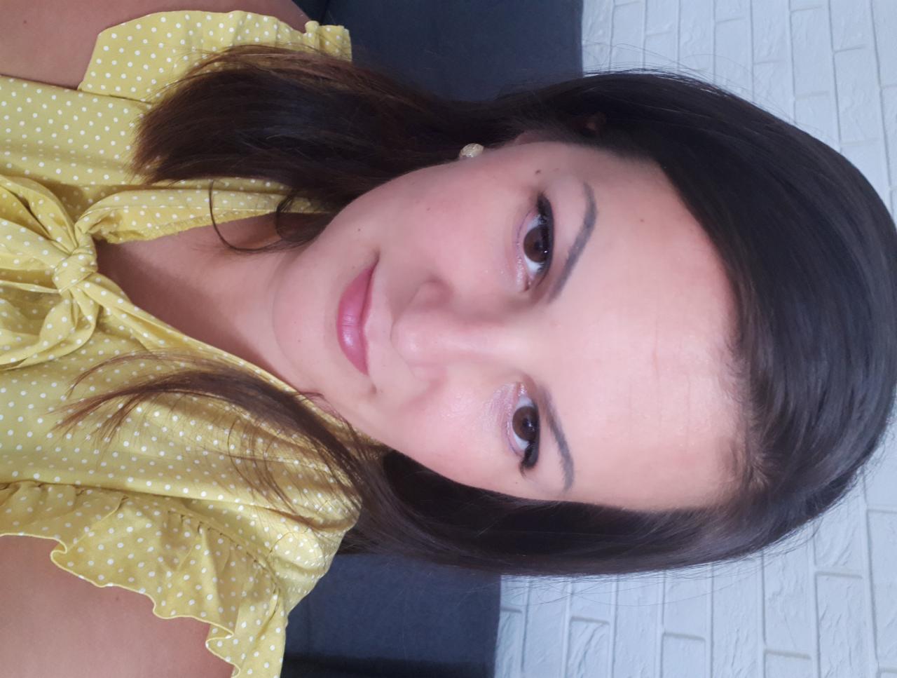 Portret uśmiechniętej kobiety w żółtej bluzce w białe kropki, z delikatnym makijażem, na tle ściany z białej cegły.