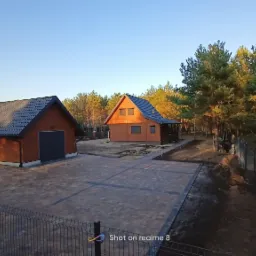 Ułożona kostka brukowa w Turek tworząca podjazd do drewnianego garażu i domu jednorodzinnego, widok zza ogrodzenia.