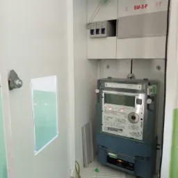 Otwarta skrzynka elektryczna z licznikiem energii i bezpiecznikami, widok z bliska na zamontowane urządzenia, przewody i oznaczenia na liczniku.